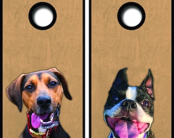May include: Two cornhole boards with dog portraits. One board features a brown and black dog with a pink collar, and the other board has a black and white dog with a blue collar. Each board has a black border and a circular hole.