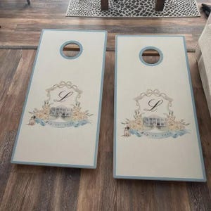 May include: Two rectangular cornhole boards with a light cream finish and light blue borders. Each board features a circular hole at the top and a central crest design containing a cursive letter L, a detailed illustration of a house, and floral accents.