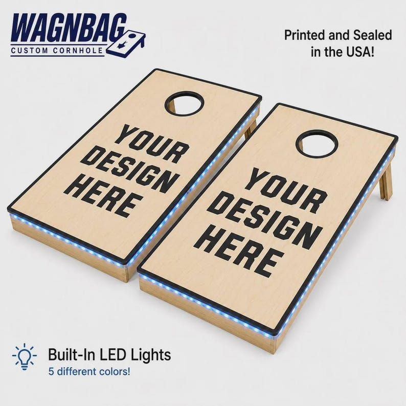 May include: Two light-colored wooden cornhole boards with black circular holes and black text reading YOUR DESIGN HERE. The boards feature integrated blue LED light strips along the edges. The WAGNBAG logo and text Printed and Sealed in the USA are visible.