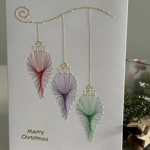 May include: A handmade Christmas card with three string-art baubles in red, purple, and green suspended from a gold wire. The card has the words "Merry Christmas" in gold lettering. The baubles are made with colourful thread.