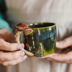 Può includere: Una tazza in ceramica fatta a mano con un'esclusiva maniglia a forma di fungo. La tazza è smaltata in tonalità verdi e blu, con un accento di fungo marrone e bianco. La tazza è tenuta in due mani.