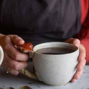May include: A handmade ceramic mug with a unique handle design. The mug is a light gray color and filled with a dark liquid. The handle features a textured, organic shape with a reddish-brown accent. The mug is held by a person wearing a red shirt and a black apron.