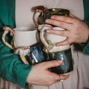 May include: A collection of ceramic mugs in shades of white, dark green, and blue. Each mug has a distinctive mushroom-shaped handle with brown and cream detailing. The mugs are held together, highlighting their handcrafted nature and unique design.