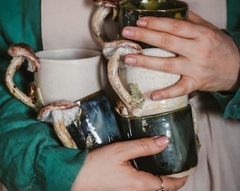 May include: A collection of ceramic mugs in shades of white, dark green, and blue. Each mug has a distinctive mushroom-shaped handle with brown and cream detailing. The mugs are held together, highlighting their handcrafted nature and unique design.