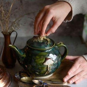 Può includere: Teiera artigianale in ceramica con smalto verde scuro. La teiera presenta un design a fungo sul lato e un manico a forma di fungo sul coperchio. La teiera è appoggiata su un vassoio di legno.