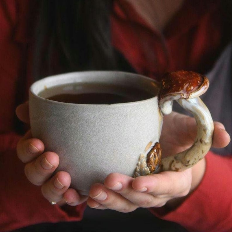 May include: A ceramic mug with a mushroom-shaped handle. The mug is light gray and filled with a dark liquid. The handle is a detailed mushroom with brown and cream tones. The mug is held in both hands.
