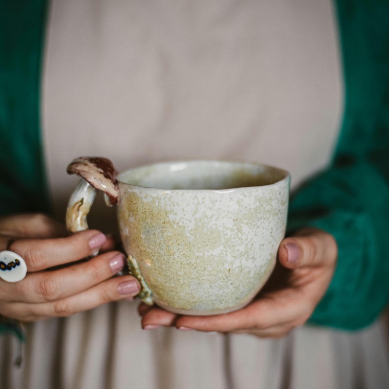 May include: A handmade ceramic mug with a mushroom-shaped handle. The mug has a speckled, off-white glaze with hints of green and brown. The mushroom handle is detailed with brown and beige tones. The mug is held in both hands.