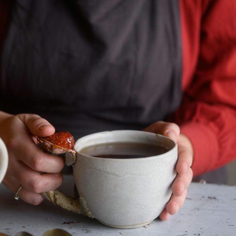 May include: A handmade ceramic mug with a unique handle design. The mug is a light gray color and filled with a dark liquid. The handle features a textured, organic shape with a reddish-brown accent. The mug is held by a person wearing a red shirt and a black apron.