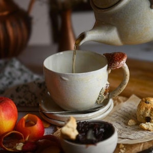 May include: A ceramic teacup and saucer set with a mushroom-shaped handle. Tea is being poured from a matching teapot. The cup is off-white with a textured finish. A small bowl of jam and a scone are also visible.