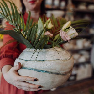 Könnte beinhalten: Eine runde, cremefarbene Keramikvase mit horizontalen blauen Streifen, gefüllt mit grünen Blättern und rosa und weißen Blumen. Die Vase hat eine strukturierte Oberfläche. Die Vase ist ein dekorativer Gegenstand.