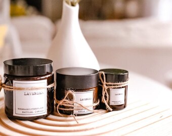 May include: Three brown glass candle jars with black lids and twine tied around the top. The jars are labeled "Lavender" and "Natural Candle". The candles are sitting on a wooden surface.