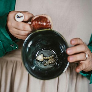 May include: A dark teal ceramic bowl with a mushroom-shaped object inside. A person holds the bowl, with a ring featuring a key design on one finger and a small, round object on another. The person wears a green shirt and a beige apron.
