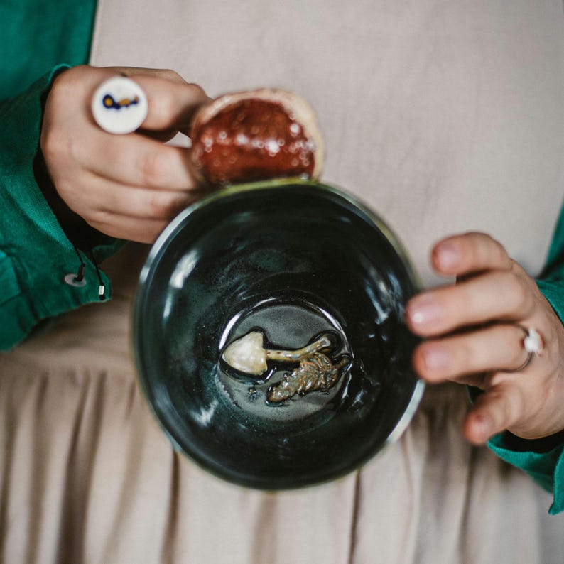 May include: A dark teal ceramic bowl with a mushroom-shaped object inside. A person holds the bowl, with a ring featuring a key design on one finger and a small, round object on another. The person wears a green shirt and a beige apron.