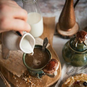 K&ouml;nnte beinhalten: Eine Keramiktasse mit einem Pilzdesign wird mit Kaffee gef&uuml;llt, w&auml;hrend Milch aus einem kleinen Krug gegossen wird. Eine Zuckerdose und eine Milchflasche sind ebenfalls zu sehen. Die Szene deutet auf eine gem&uuml;tliche, rustikale &Auml;sthetik hin.