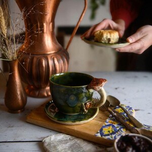 May include: A ceramic teacup and saucer set with a mushroom-shaped handle and accents. The cup is dark green with blue and brown details, sitting on a wooden board with a spoon and a small plate. A copper pitcher and vase are in the background.