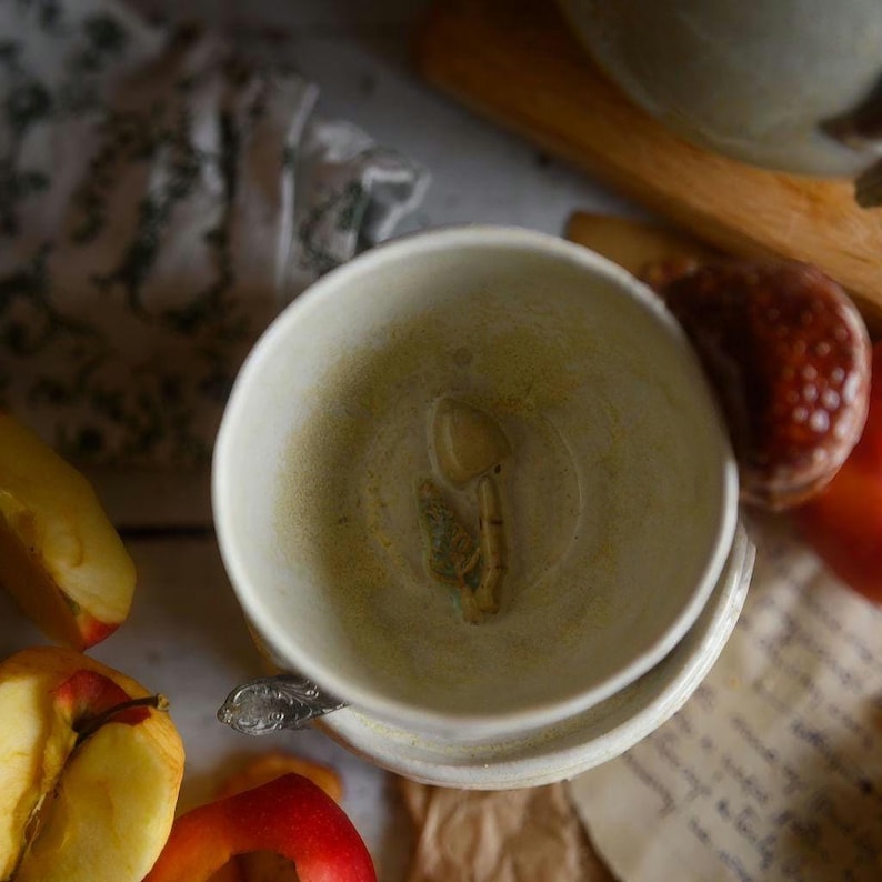 May include: A white ceramic teacup with a mushroom design inside, resting on a saucer. The cup has a light green interior and is surrounded by apple slices and a vintage spoon. The scene evokes a rustic, cozy atmosphere.