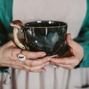 May include: A ceramic mug with a dark teal glaze and a mushroom-shaped handle. The mug has a wide, rounded shape and a cream-colored interior. The mushroom handle is brown and beige. The mug is held in both hands.