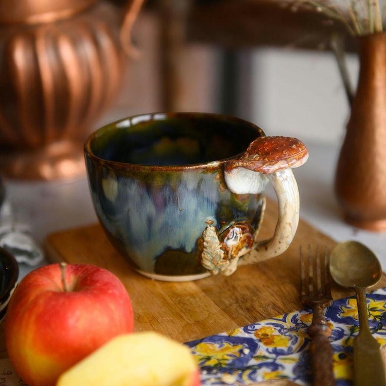 May include: A ceramic mug with a mushroom handle and a mushroom accent on the side. The mug has a blue and green glaze with brown accents. The mug is sitting on a wooden cutting board with an apple in the foreground.