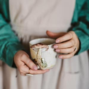K&ouml;nnte beinhalten: Eine Keramiktasse mit einem Pilzdesign. Die Tasse ist cremefarben mit einem braunen und beigen Pilzakzent an der Seite. Die Tasse wird von einer Person gehalten, die eine gr&uuml;ne Jacke und eine beige Sch&uuml;rze tr&auml;gt.