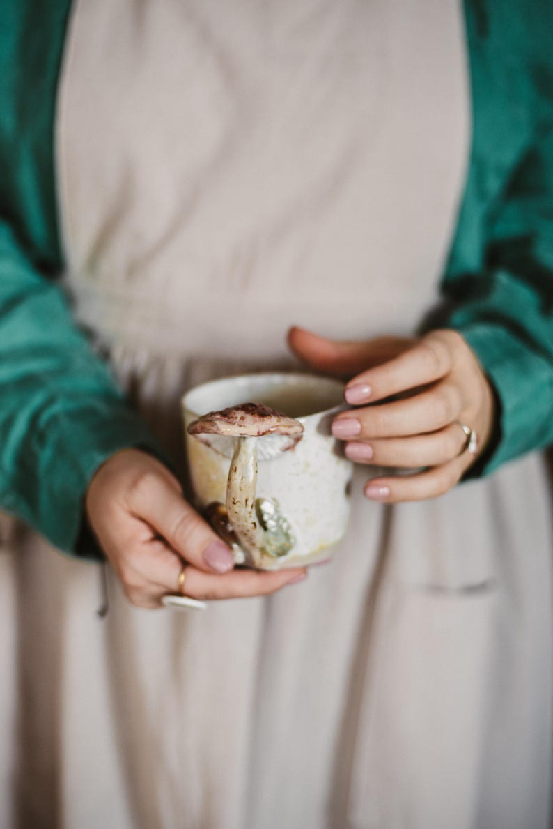 K&ouml;nnte beinhalten: Eine Keramiktasse mit einem Pilzdesign. Die Tasse ist cremefarben mit einem braunen und beigen Pilzakzent an der Seite. Die Tasse wird von einer Person gehalten, die eine gr&uuml;ne Jacke und eine beige Sch&uuml;rze tr&auml;gt.
