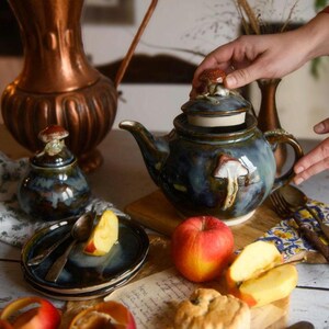 May include: A ceramic teapot and sugar bowl set with mushroom accents. The teapot is dark blue with a mushroom design and a mushroom-shaped lid handle. The sugar bowl has a similar design. Apples and plates with spoons are also visible.