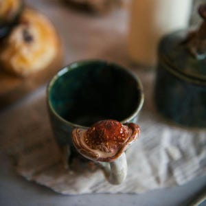 K&ouml;nnte beinhalten: Eine handgefertigte Keramik-Tasse mit einem pilzf&ouml;rmigen Griff. Die Tasse ist dunkelblaugr&uuml;n mit gl&auml;nzendem Finish, und der Pilzgriff weist Braun- und Cremet&ouml;ne auf. Die Tasse steht auf einer strukturierten Oberfl&auml;che.