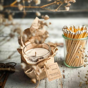 Könnte beinhalten: Eine in braunes Papier gewickelte Kerze in einem durchsichtigen Glasgefäß mit braunem Deckel. Die Kerze ist mit "Coffee" beschriftet und hat ein braunes, natürliches Wachs. Die Kerze steht auf einer weißen Holzoberfläche mit einem Streichholzschachtel im Hintergrund.