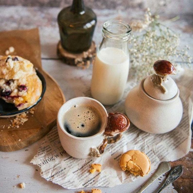 K&ouml;nnte beinhalten: Ein Stillleben mit einer Kaffeetasse, einer Milchflasche und einer Zuckerdose mit pilzf&ouml;rmigem Deckel. Ein Muffin und ein Macaron sind ebenfalls vorhanden. Die Gegenst&auml;nde sind auf einer Zeitung und einer Holzoberfl&auml;che angeordnet.