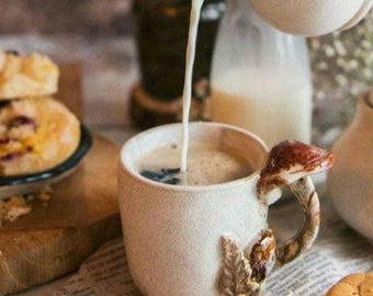 May include: A cream-colored ceramic mug with a mushroom-shaped handle and leaf accents. Coffee is being poured into the mug from a matching pitcher. Baked goods are visible in the background.