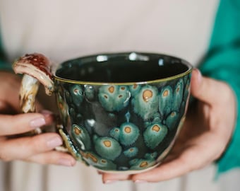 May include: A ceramic mug with a dark green and teal glaze, featuring a mushroom-shaped handle. The mug has a speckled pattern with orange accents. The handle is brown and beige. The mug is held by a person.