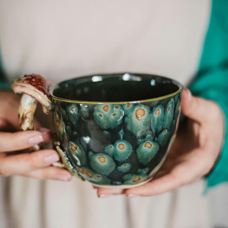 May include: A ceramic mug with a dark green and teal glaze, featuring a mushroom-shaped handle. The mug has a speckled pattern with orange accents. The handle is brown and beige. The mug is held by a person.