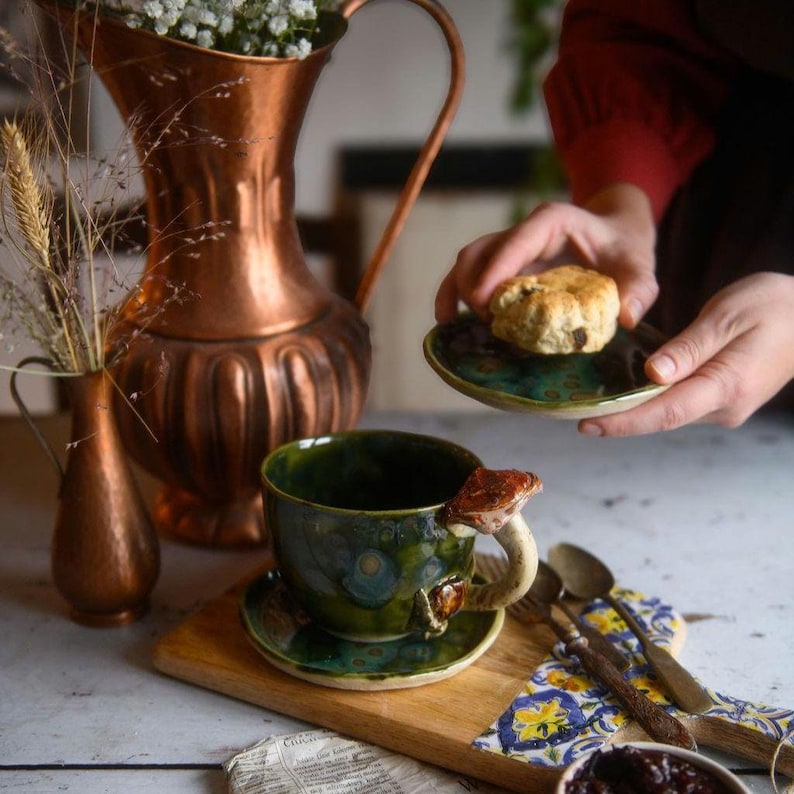 May include: A handmade ceramic teacup and saucer set with a mushroom handle. The cup and saucer are glazed in shades of green and blue. A copper pitcher and vase are in the background, along with a wooden cutting board and a small plate with a scone.