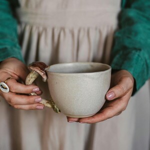 May include: A beige ceramic mug with a mushroom-shaped handle. The mushroom handle has a brown cap and a beige stem. The mug is held by two hands with pink nail polish. The mug is a unique and whimsical addition to any kitchen.