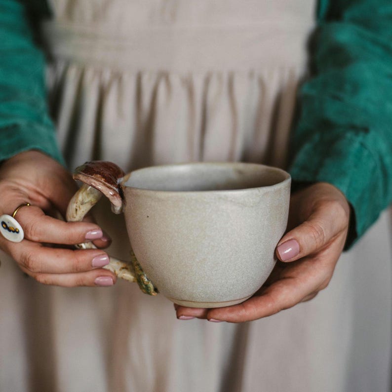 May include: A beige ceramic mug with a mushroom-shaped handle. The mushroom handle has a brown cap and a beige stem. The mug is held by two hands with pink nail polish. The mug is a unique and whimsical addition to any kitchen.