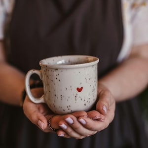 Könnte beinhalten: Eine gesprenkelte, weiße Keramiktasse mit einem roten Herz. Die Tasse hat eine abgerundete Form und einen Henkel. Die Tasse wird in beiden Händen gehalten. Es ist eine Kaffeetasse.
