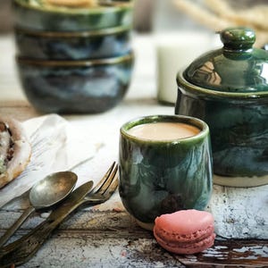 May include: A close-up of a green ceramic cup filled with a light-coloured beverage, a pink macaron, and antique silverware. In the background are stacked bowls and a lidded jar, all in shades of green and blue.