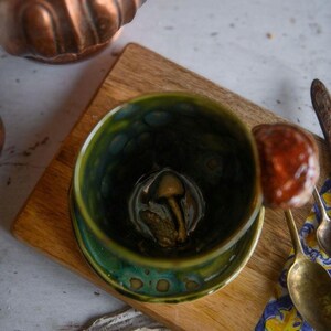 May include: A handmade ceramic bowl with a green and blue glaze, featuring a mushroom-shaped object inside. The bowl sits on a wooden board, with a copper-colored bowl in the background. A spoon with a mushroom is on the right.