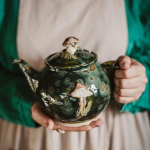 Może przedstawiać: Ręcznie wykonany ceramiczny imbryk z zielono-brązową glazurą. Imbryk posiada uchwyt w kształcie grzyba na pokrywce oraz aplikację grzyba na korpusie. Imbryk jest trzymany na tle neutralnego tła.