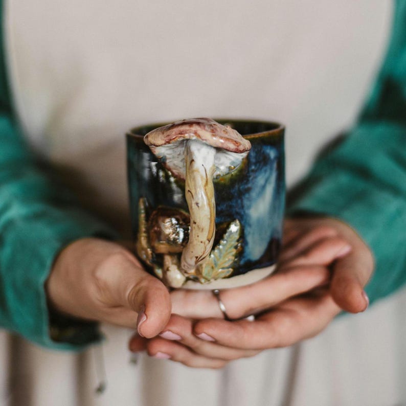 K&ouml;nnte beinhalten: Eine Keramiktasse mit einem Pilzgriff und dekorativen Pilz- und Bl&auml;ttermotiven. Die Tasse hat eine dunkelblaue und gr&uuml;ne Glasur mit einem braunen und wei&szlig;en Pilzgriff. Die Tasse wird in zwei H&auml;nden gehalten.