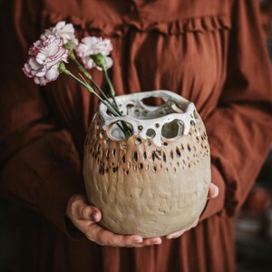 May include: A handmade ceramic vase with a distinctive design. The vase features a blend of beige and brown hues, a textured finish, and numerous holes around the midsection. It contains several pink and white carnations, forming a natural, artistic arrangement.