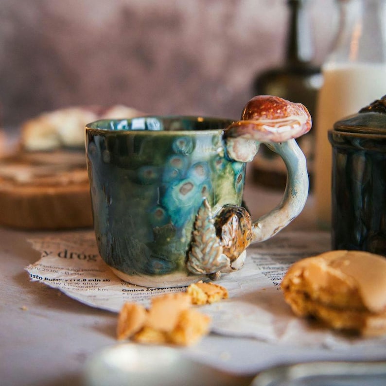 K&ouml;nnte beinhalten: Eine Keramik-Tasse mit einem Pilzgriff und Akzenten. Die Tasse ist in T&uuml;rkis- und Gr&uuml;nt&ouml;nen glasiert, mit einem braunen und roten Pilz am Griff. Die Tasse steht auf einer Zeitung.