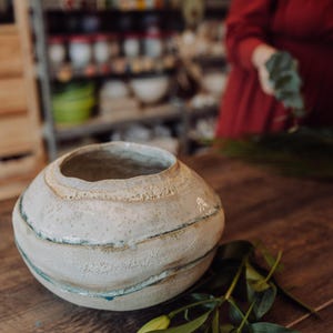 Pode incluir: Um vaso redondo de cerâmica esbranquiçada com uma abertura larga. O vaso tem uma superfície texturizada com faixas horizontais de cor, incluindo azul claro e bege. O vaso está sobre uma superfície de madeira.