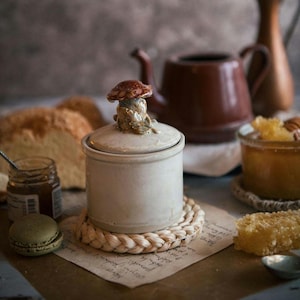 Può includere: Un piccolo barattolo di ceramica bianco sporco con un coperchio a forma di fungo. Il barattolo è posto su un sottobicchiere intrecciato, con un piccolo barattolo di marmellata, un macaron e un pezzo di favo nelle vicinanze. Una teiera marrone e del pane sullo sfondo.