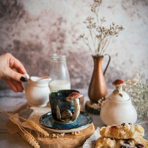 K&ouml;nnte beinhalten: Ein Keramik-Teebecher und Untertasse mit einem Pilzgriff und Deckel. Die Tasse ist dunkelblau mit einem braunen Pilzgriff. Eine kleine wei&szlig;e Sahnek&auml;nnchen und eine Milchflasche sind ebenfalls vorhanden, zusammen mit einer kleinen Vase mit Trockenblumen.
