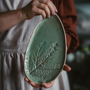 May include: A handmade, oval ceramic serving platter in a speckled teal hue. It features a raised, textured design of a pine branch. The rim is a lighter, natural clay colour. The platter is held by a person.