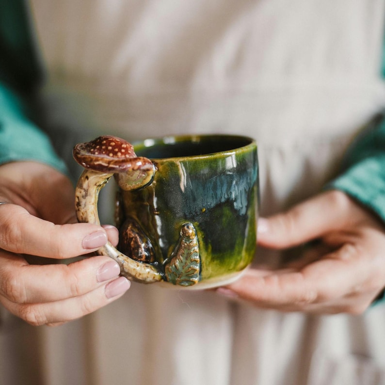 K&ouml;nnte beinhalten: Eine handgefertigte Keramiktasse mit einem einzigartigen Pilzgriff. Die Tasse ist in Gr&uuml;n- und Blaut&ouml;nen glasiert, mit einem braunen und wei&szlig;en Pilzakzent. Die Tasse wird in zwei H&auml;nden gehalten.