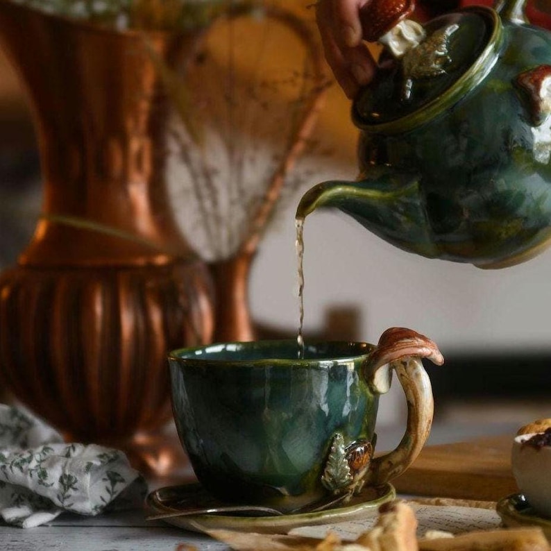 May include: A dark green ceramic teacup and saucer set with a mushroom-shaped handle and leaf accents. A matching teapot pours tea into the cup. The teapot lid is decorated with a mushroom and strawberry.