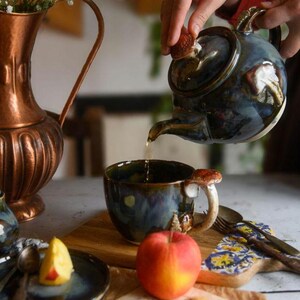 May include: A blue and brown ceramic teapot pouring tea into a matching mug. The mug and teapot feature mushroom-shaped details. A copper pitcher and an apple are also visible on the wooden surface.