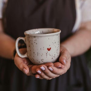 Könnte beinhalten: Eine gesprenkelte weiße Keramik-Tasse mit einem roten Herz-Design. Die Tasse hat eine abgerundete Form, einen Henkel und ist mit einer dunklen Flüssigkeit gefüllt. Die Tasse wird mit beiden Händen gehalten.
