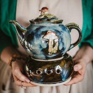 Könnte beinhalten: Keramik-Teekanne mit blau-brauner Glasur, mit einem Pilzdesign auf Körper und Deckel. Die Teekanne steht auf einem passenden Wärmer mit drei kreisförmigen Öffnungen.